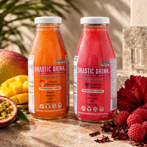 Drastic Drink Raspberry Hibiscus and Mango Passionfruit outside beside hibiscus flower, raspberries, whole mango, cut mango, cut passionfruit, with plant in the background.