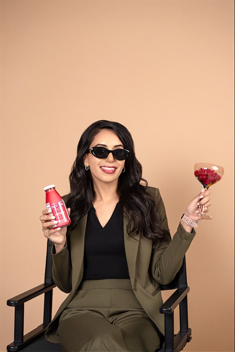 A woman (Founder Simmy Sidhu) wearing a matching olive green blazer and trousers set with a black top sits in a black chair against a solid beige background. She is holding a dark pink Drastic Drink bottle in one hand and a cocktail glass in the other with raspberries in it.