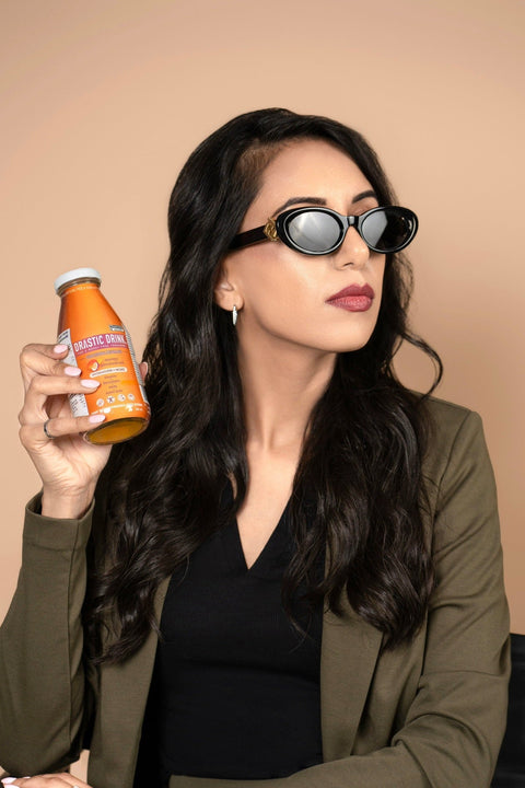 A woman (Founder Simmy Sidhu) with long dark wavy hair wearing black sunglasses and an olive green blazer holds up an orange bottle of 'Drastic Drink Mango Passionfruit' against a solid beige background.